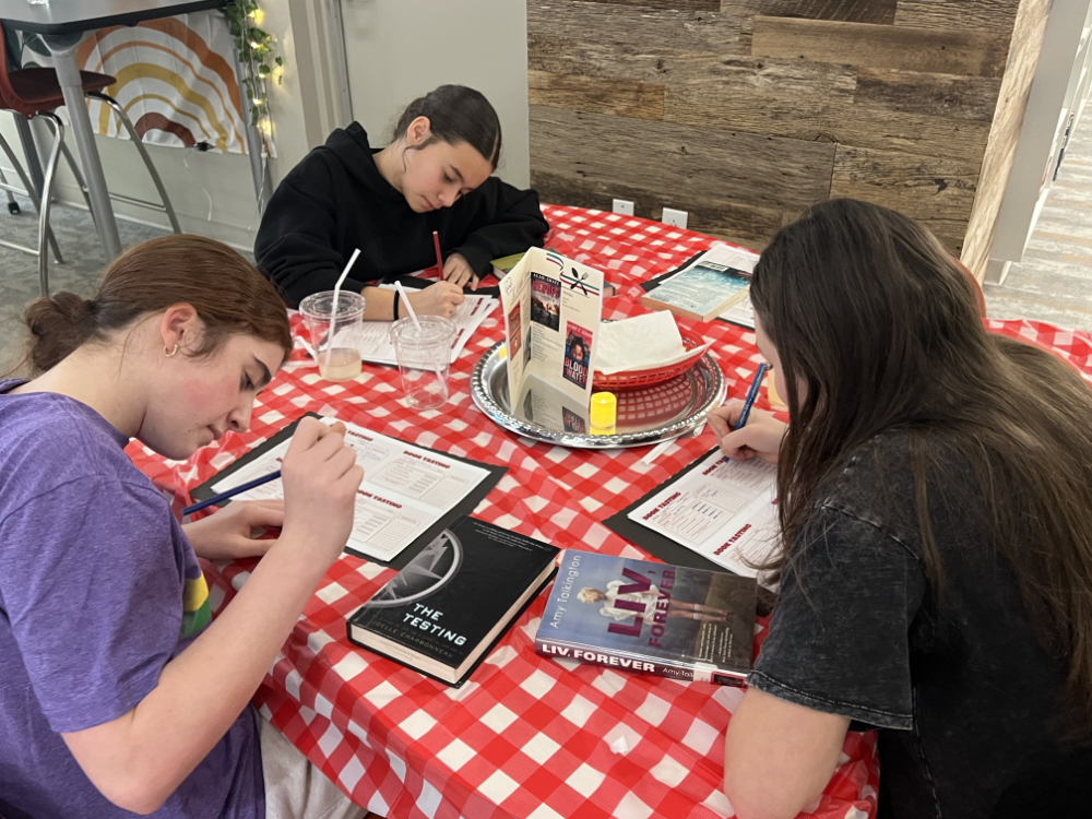 Middle school students sit at decorated tables in the Innovation Center, transformed into the IC Cafe, sampling books, taking notes, and enjoying drinks while exploring a variety of high interest titles.