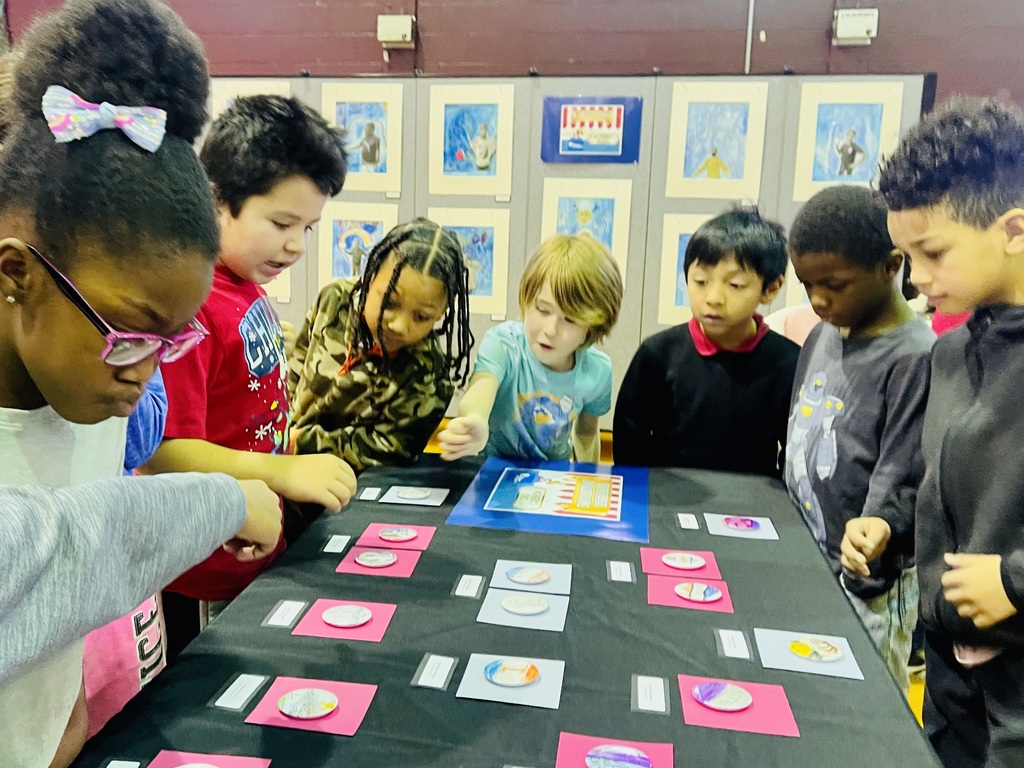 Elementary students gather around a table during a hands-on classroom activity, examining artwork or learning materials displayed on cards. The students lean in with curiosity and focus, collaborating as they explore and discuss the materials together.