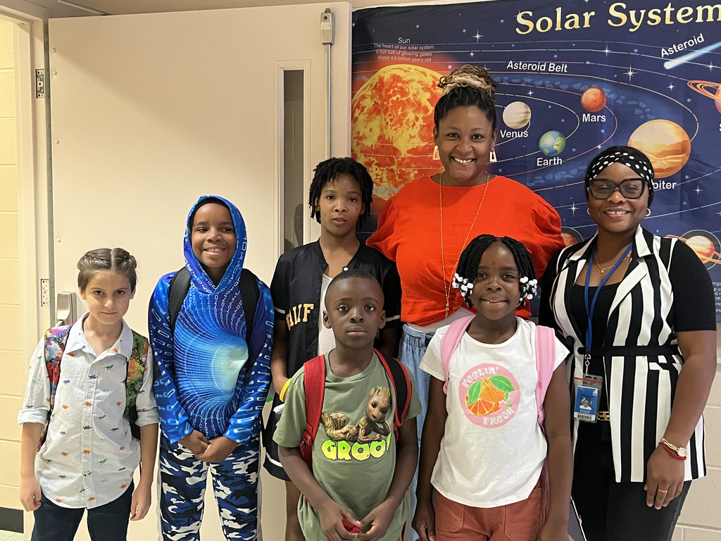 A group of elementary students stand with two adults in a school hallway, smiling in front of a colorful solar system display. The students wear backpacks and casual clothing, representing a diverse group of learners at Stratton Academy of the Arts.