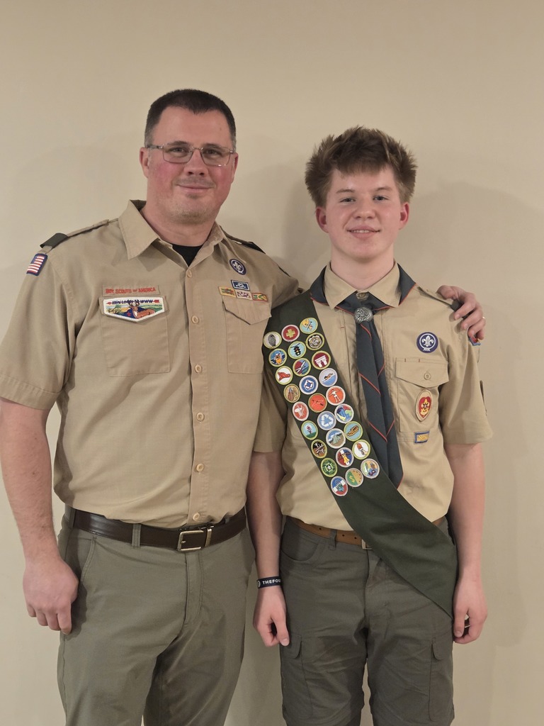 Two men in tan uniforms. One is wearing glasses, a belt, and has a sash with badges.
