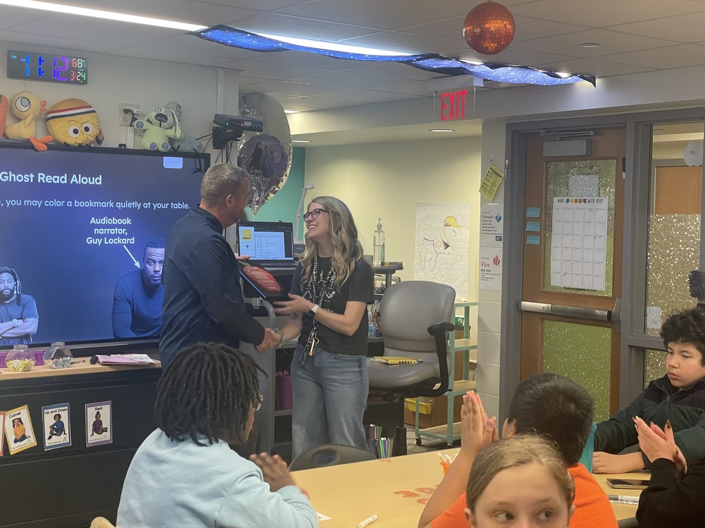 Classroom with a large screen. A man and a woman are standing in front of a screen, presenting. Students sit at desks, clapping.