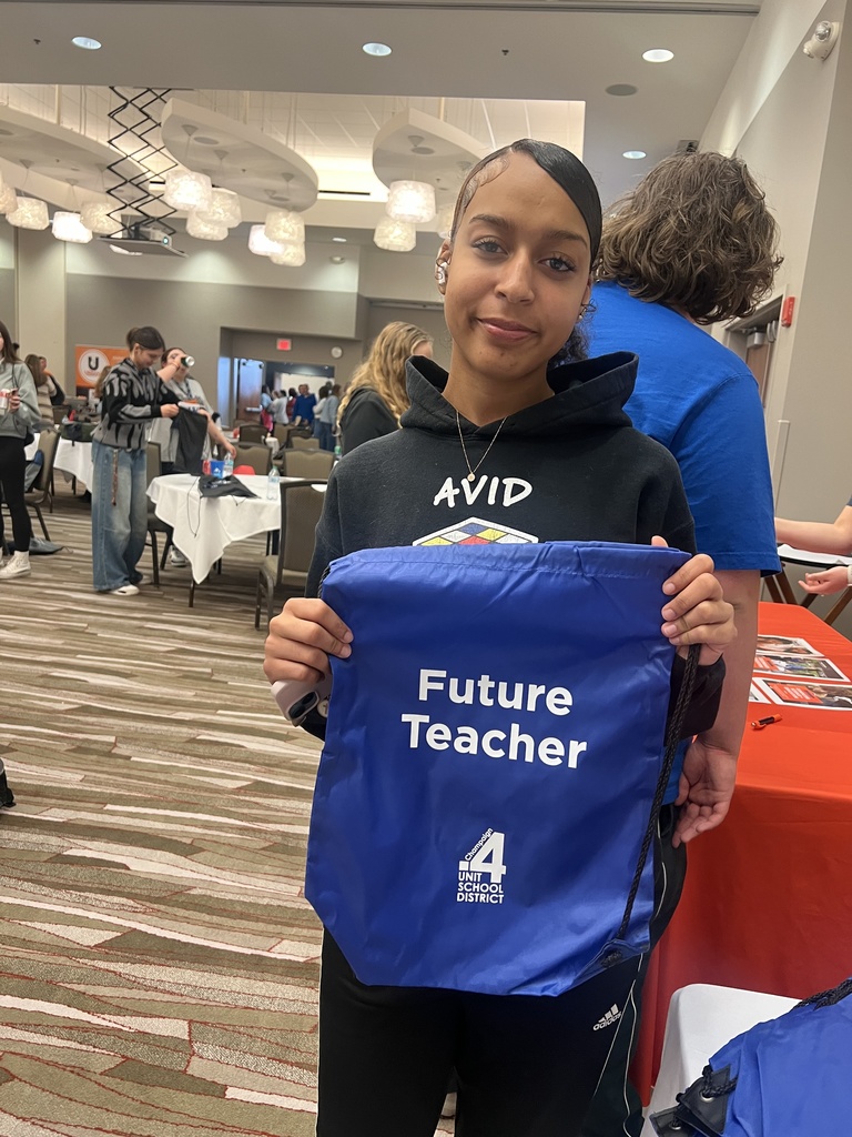 Woman holds a blue bag reading "Future Teacher" with a logo and number 4. She is at an event with people and tables in the background.