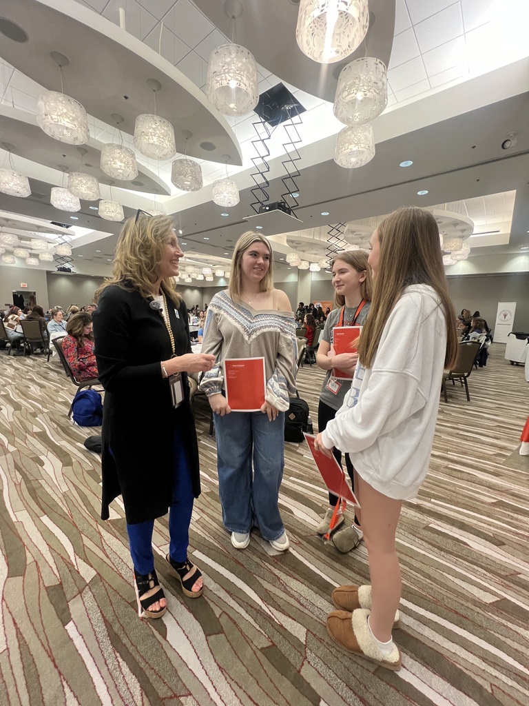 Three women holding red folders stand in a hall with a striped floor, facing each other. Ceiling lights hang above.