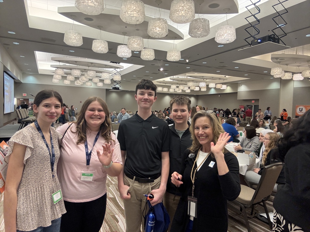 A group of people with name tags stands in a conference room, smiling at the camera. The room is filled with people seated and standing.