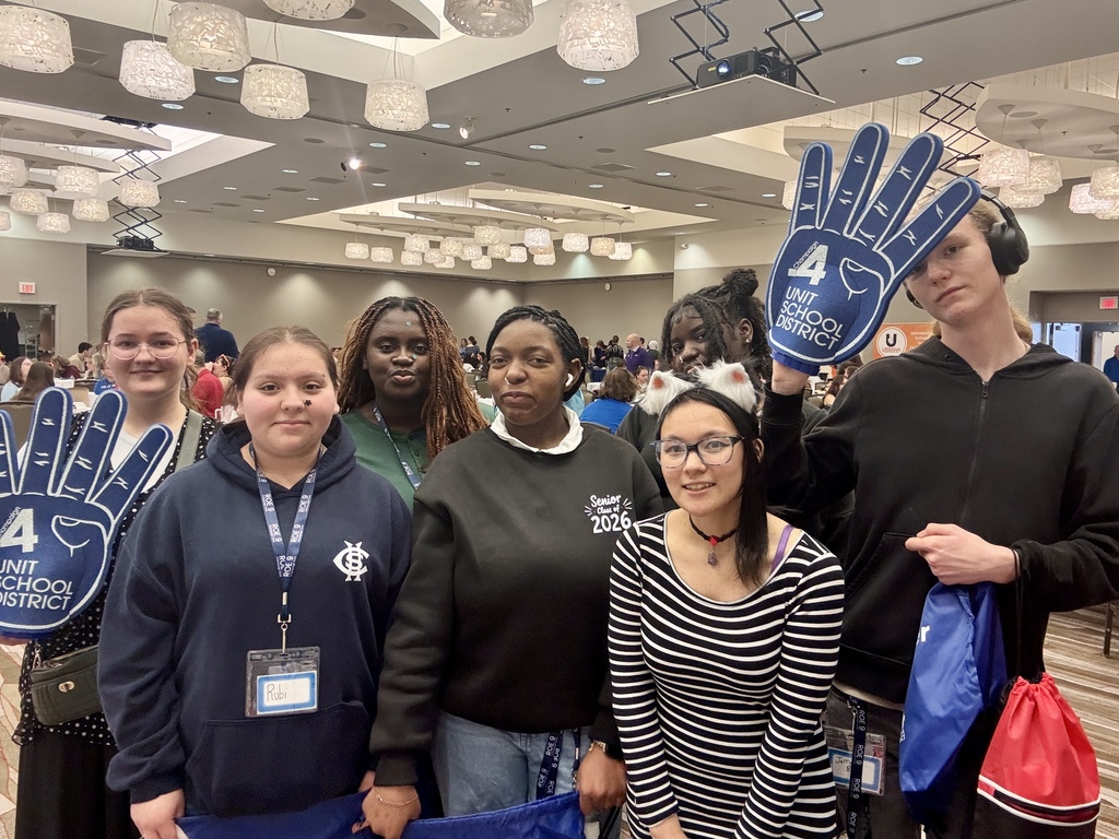 Six people stand in a room with hands holding up signs. They wear jackets and IDs. A woman has glasses.
