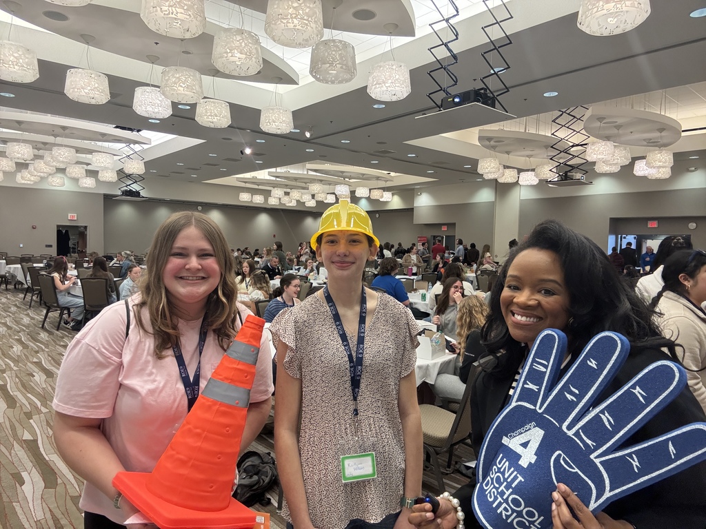 Three women in a large room with many people in the background. They hold a traffic cone, a helmet, and a glove.