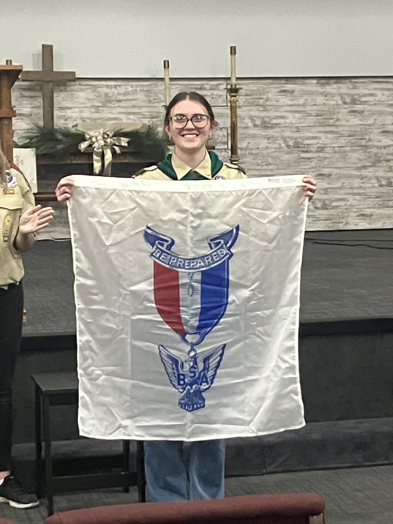 A young person holds a flag with a crest, in a church setting. Another person stands beside them.
