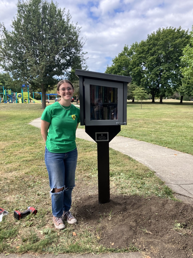 A woman stands next to a black box on a post, holding a book. Behind her, a paved pathway leads to playground equipment.