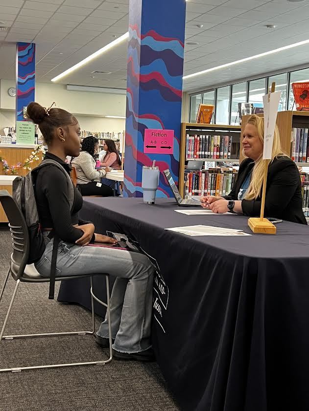 A person sits at a table with another person in a library. They have a laptop in front of them.