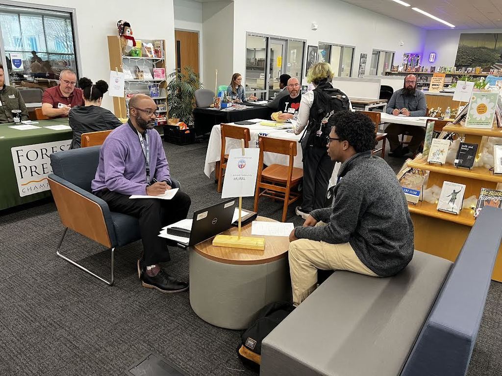 People seated in chairs in a room with tables and shelves filled with books. One man looks at a laptop.