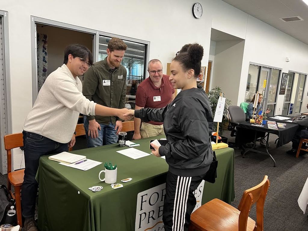 People stand around a table with a green cover and a banner reading "FOR PRE". A woman shakes hands with a man.