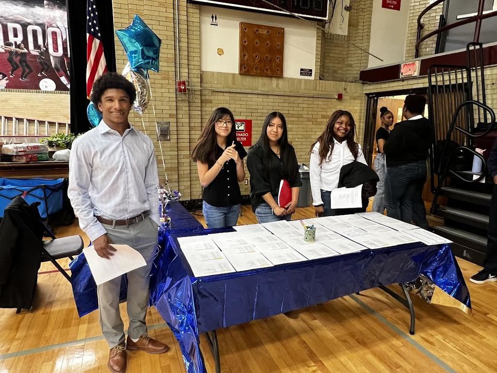 Five people stand at a table covered in blue, holding papers and a cup. A flag and balloons are behind them.