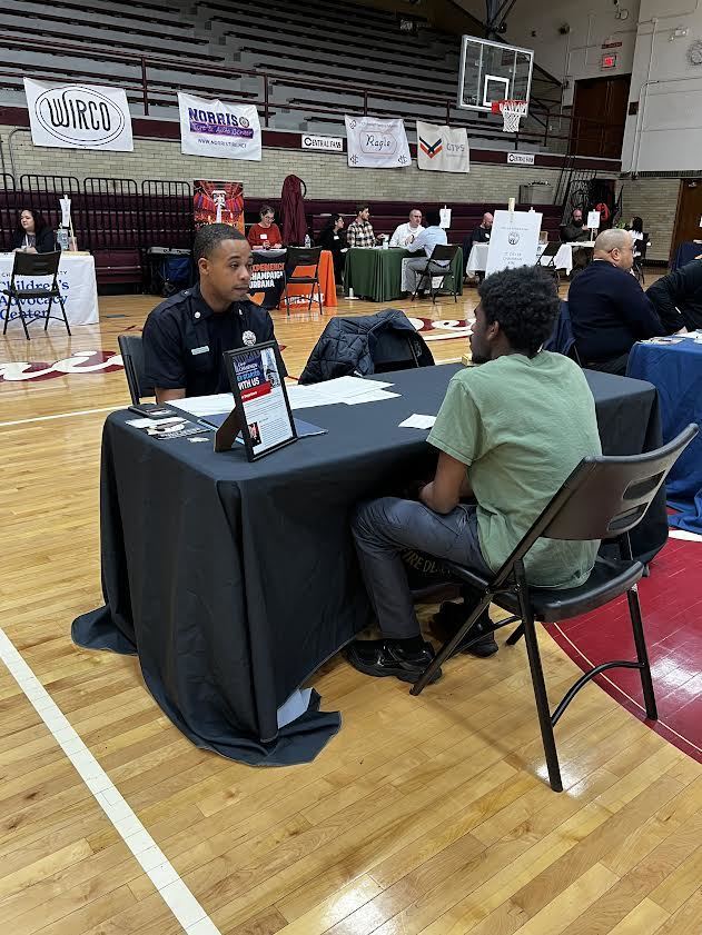 Two men sit at a table in a gymnasium. One wears a green shirt, the other a police uniform.