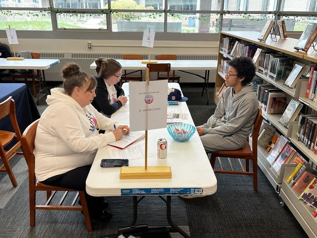 People seated at a table in a library with books on shelves behind them. One person is writing on paper.