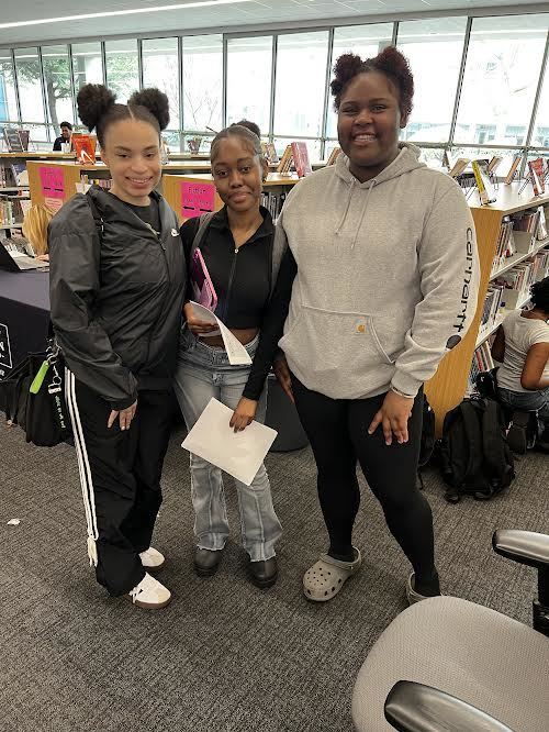 Three women in a library, with two holding papers and a backpack nearby. One wears a gray hoodie, another a black jacket.