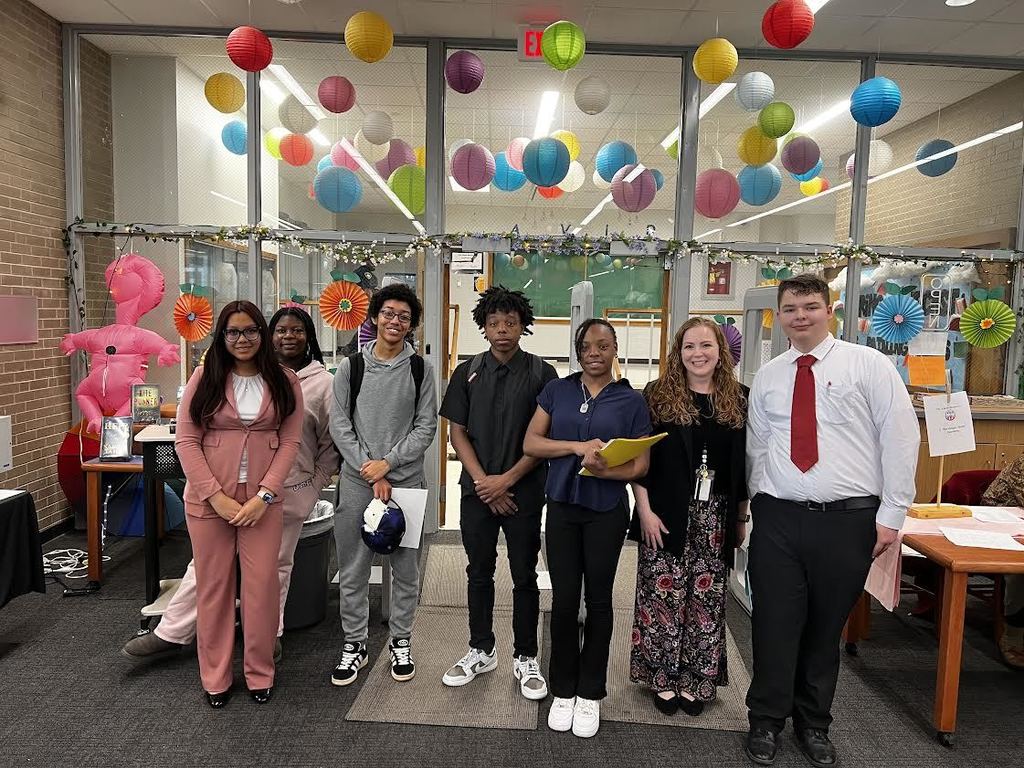 Several students and an adult male stand in a room with balloons and colorful decorations.
