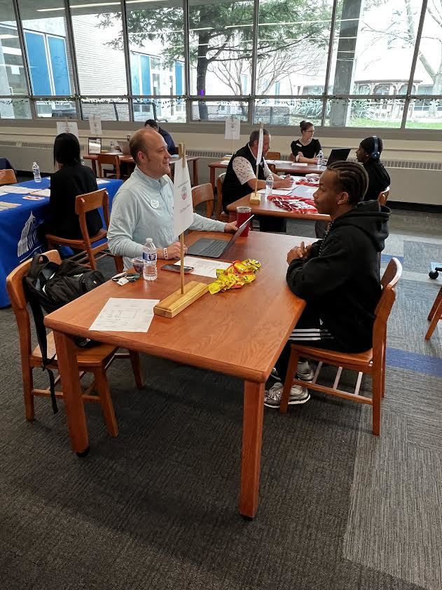A group of people are seated at desks in a room with large windows, engaged in a discussion.