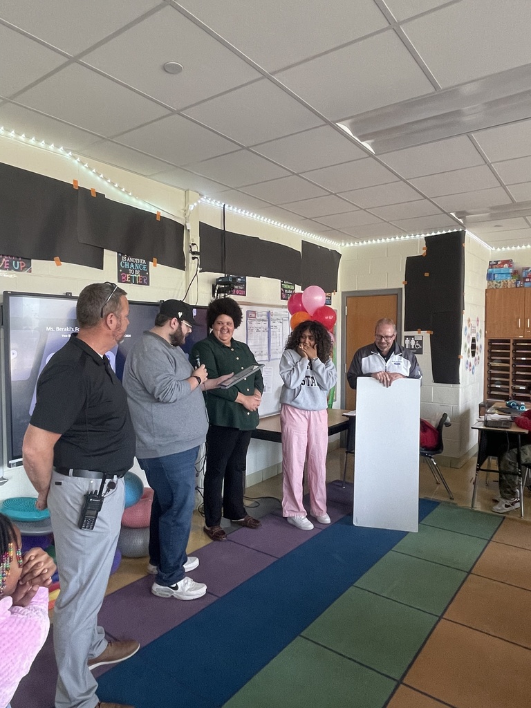A group of people gather around a podium. One person stands at the podium, others behind, and a multicolored floor mat.