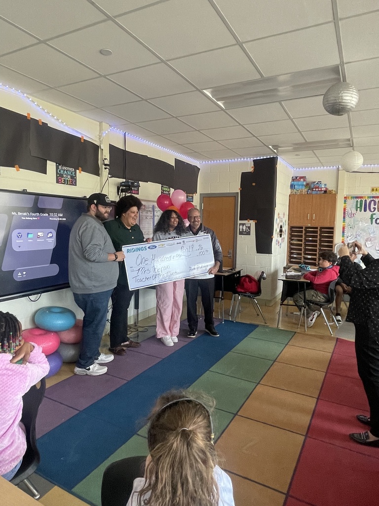 Several adults in a classroom holding a large check while a crowd of children watch.