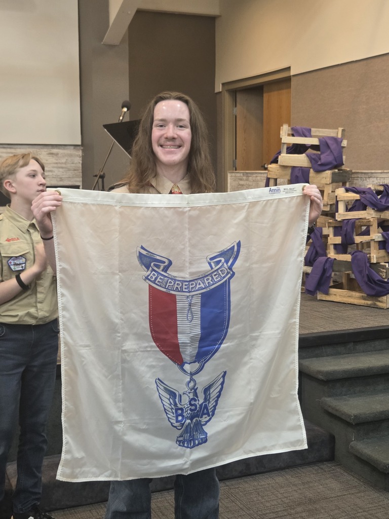 A person holds a white flag with a blue eagle emblem. Another person stands behind, both wearing uniforms.