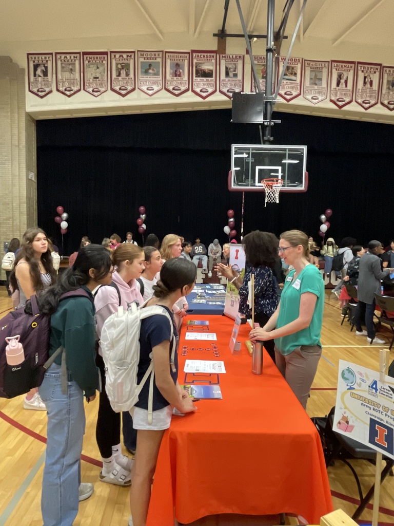 A group of students stands at a long orange table inside a gymnasium, attending an event.