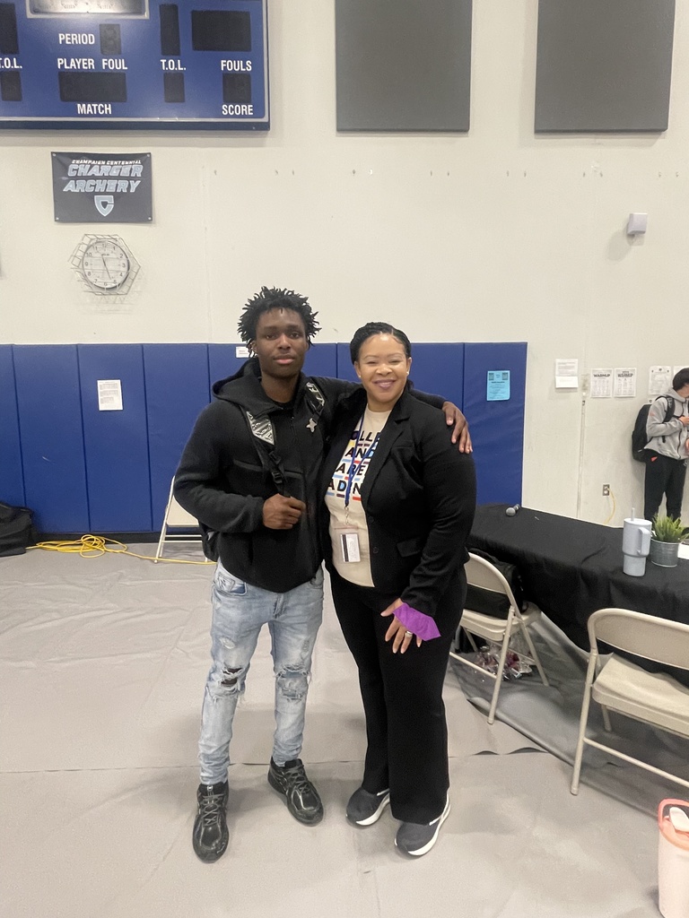 Two people standing in a gym, one with a backpack, next to a scoreboard and tables.