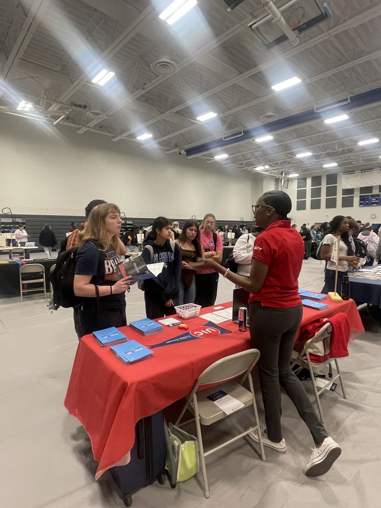 People gather around tables in a room with ceiling lights and tables covered in red cloth.