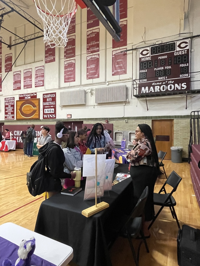 People in a basketball court stand at tables. One woman speaks to a group. A basketball hoop hangs on the wall.