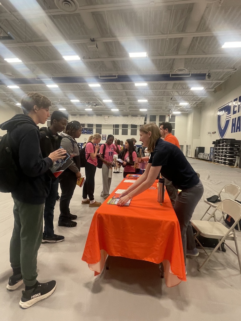 People gather around an orange table in a gymnasium. A woman interacts with them while standing.