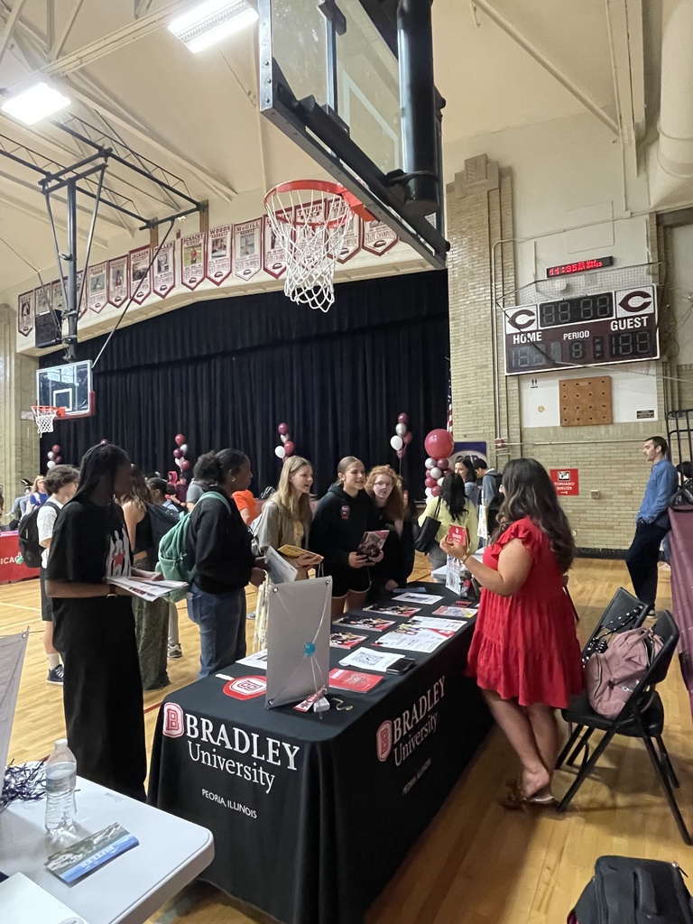 People gather in a gym, with tables and chairs. One table has a sign reading "BRADLEY UNIVERSITY".
