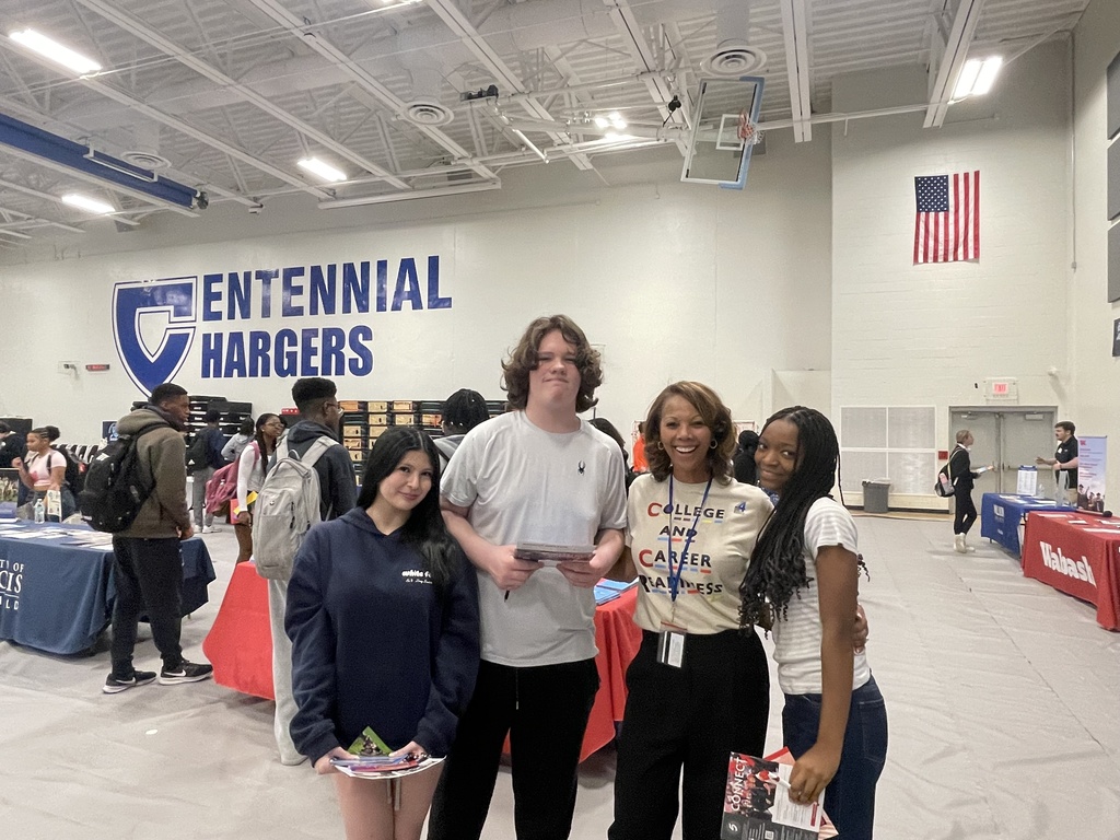 Several people are standing in a room with a large sign that reads "Enennial Hargers." Tables with displays are behind them.