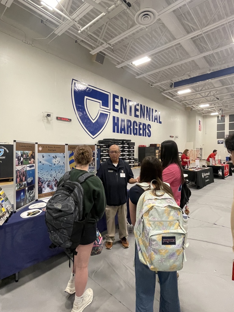 People in a room with an event banner behind them. They wear backpacks and converse. Tables are in the background.