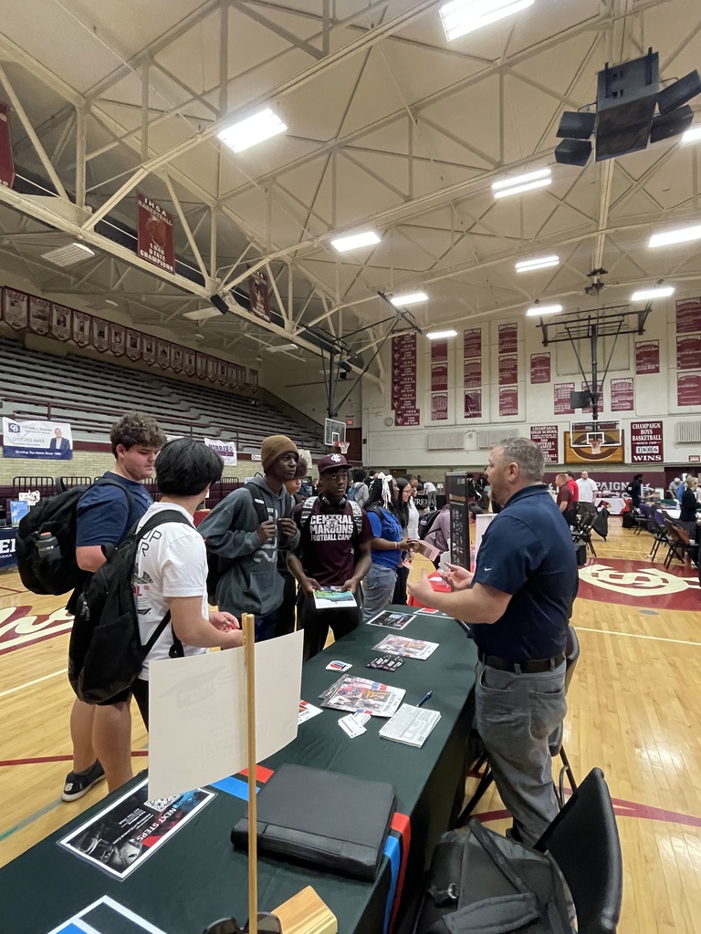 People in a gym with a basketball court; tables with materials are set up in the center.