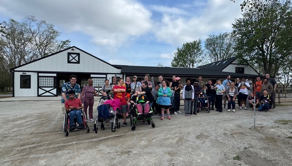 A group of people in various clothing stand in front of a barn. Some use wheelchairs.