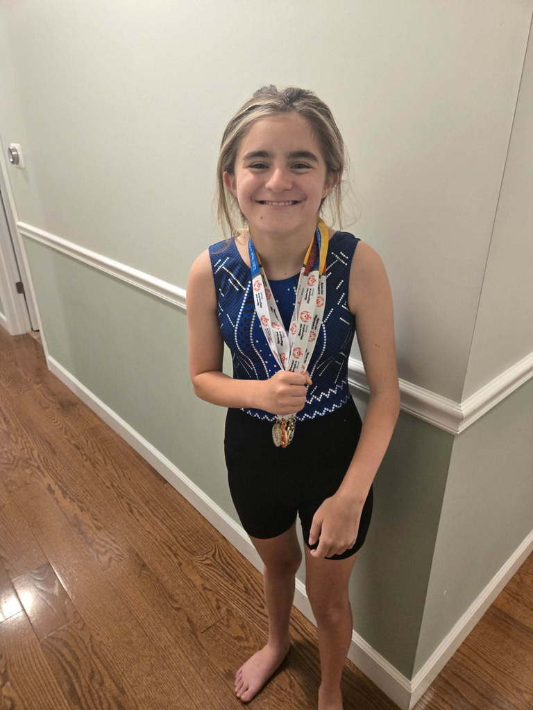 A young girl in sports gear, smiling and showing medals, stands in a hallway with wooden floors and white walls.