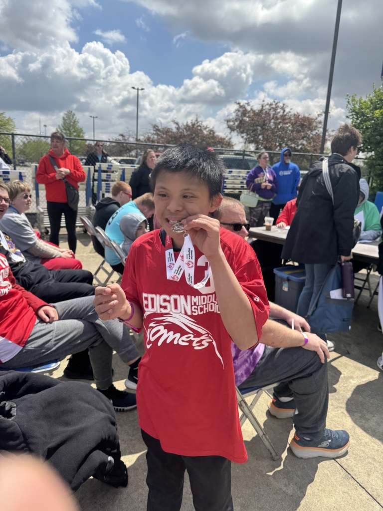 A boy wearing a red shirt poses with a medal in his mouth in a park.
