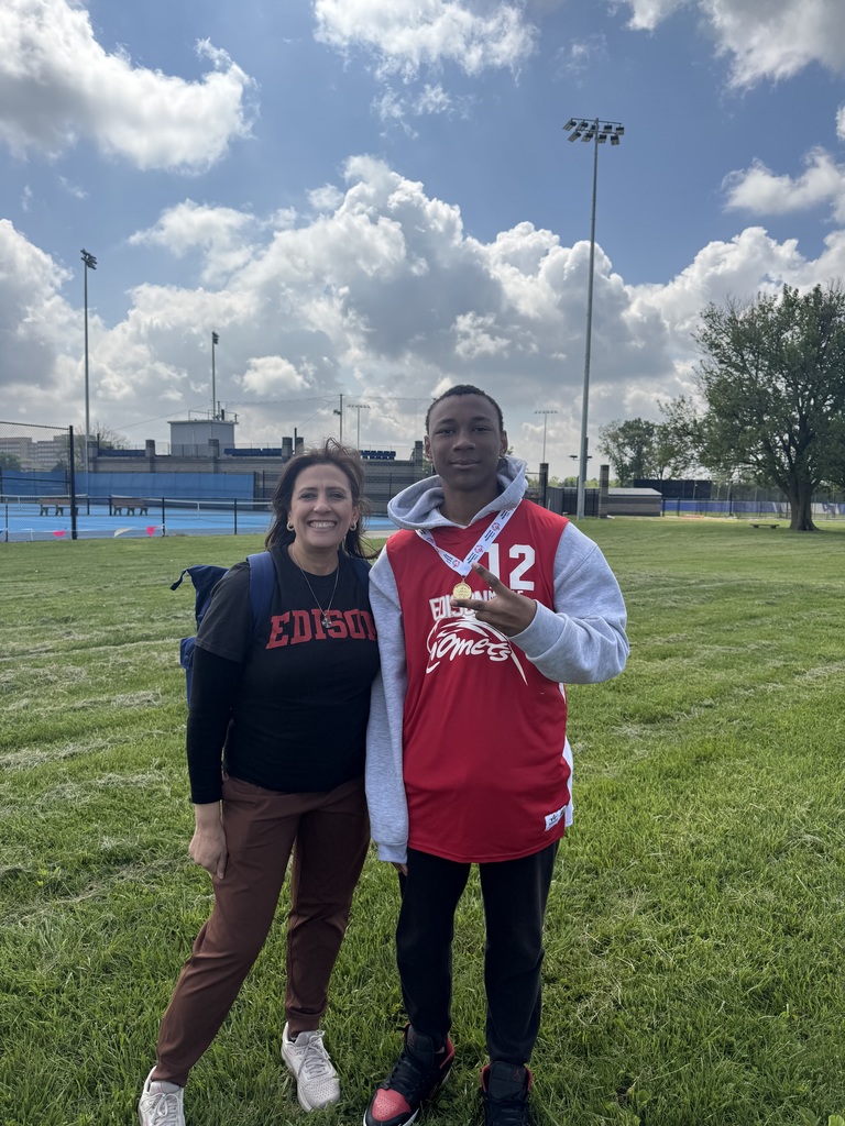 Two people stand on a grassy field. A woman in a black shirt stands next to a man in a red hoodie.