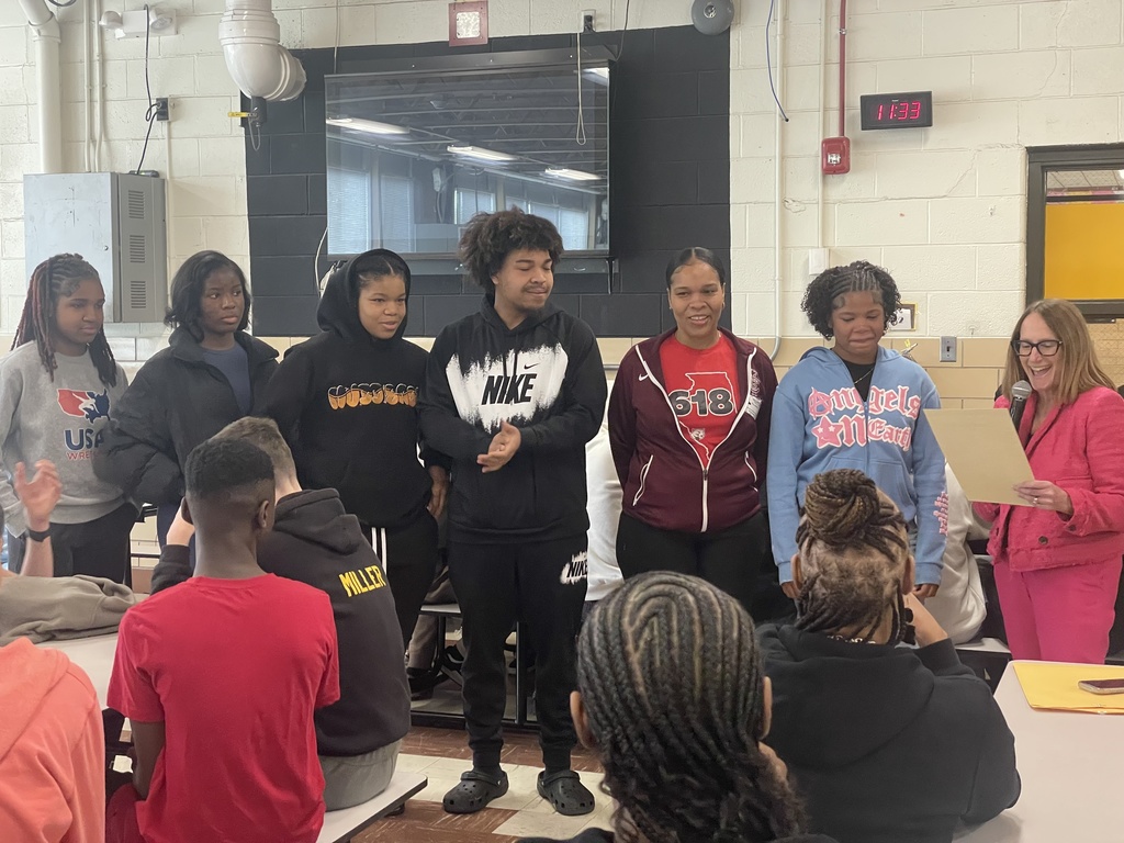 A group of students stand together in a classroom, some wearing jackets. A woman holds a clipboard.