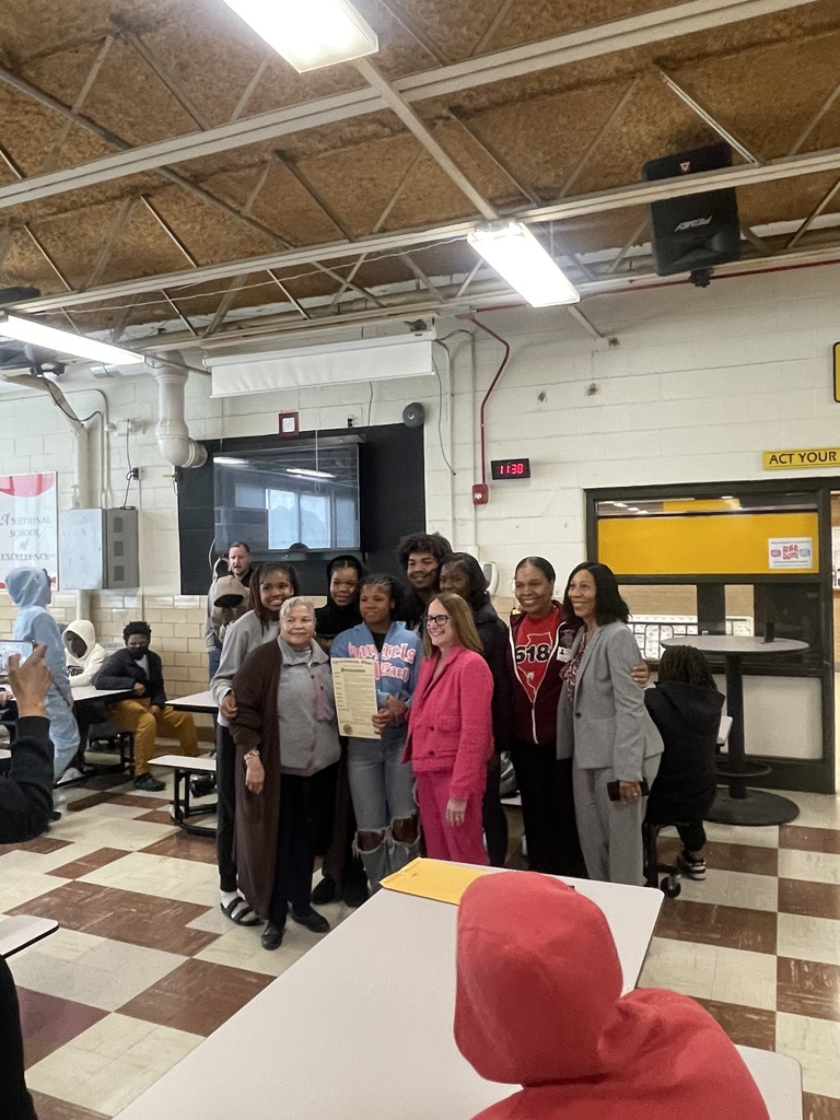 A group of women and girls in a cafeteria. One holds a certificate. A few are seated at tables.