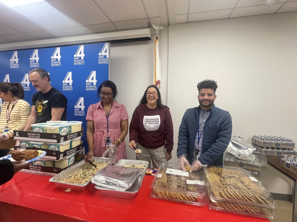 Five people stand at a table with food, including desserts. A blue banner with white numbers is behind them.