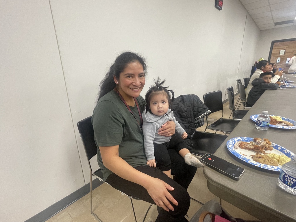 A woman holds a child on her lap, seated in a dining area. They are next to a table with plates of food.