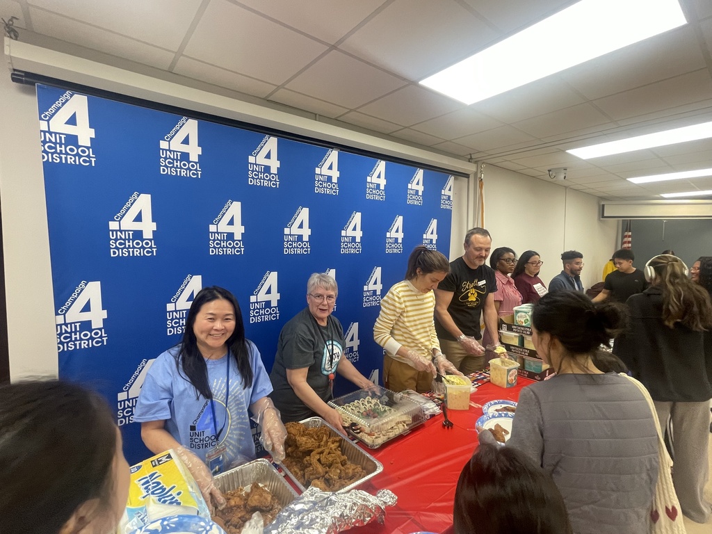 People gather around a table filled with food at an event. A blue banner with the number "4" prominently displayed in the background.