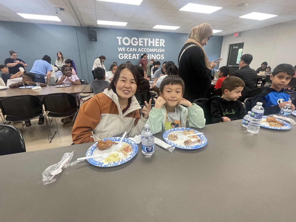 A woman and a child sit at a table with plates of food, a bottle of water, and napkins. Behind them, people are seated at tables.