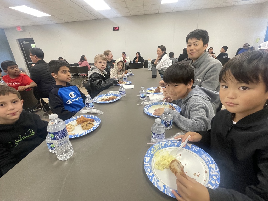 A group of children eating lunch at a table with plates, bottles, and utensils in a room.