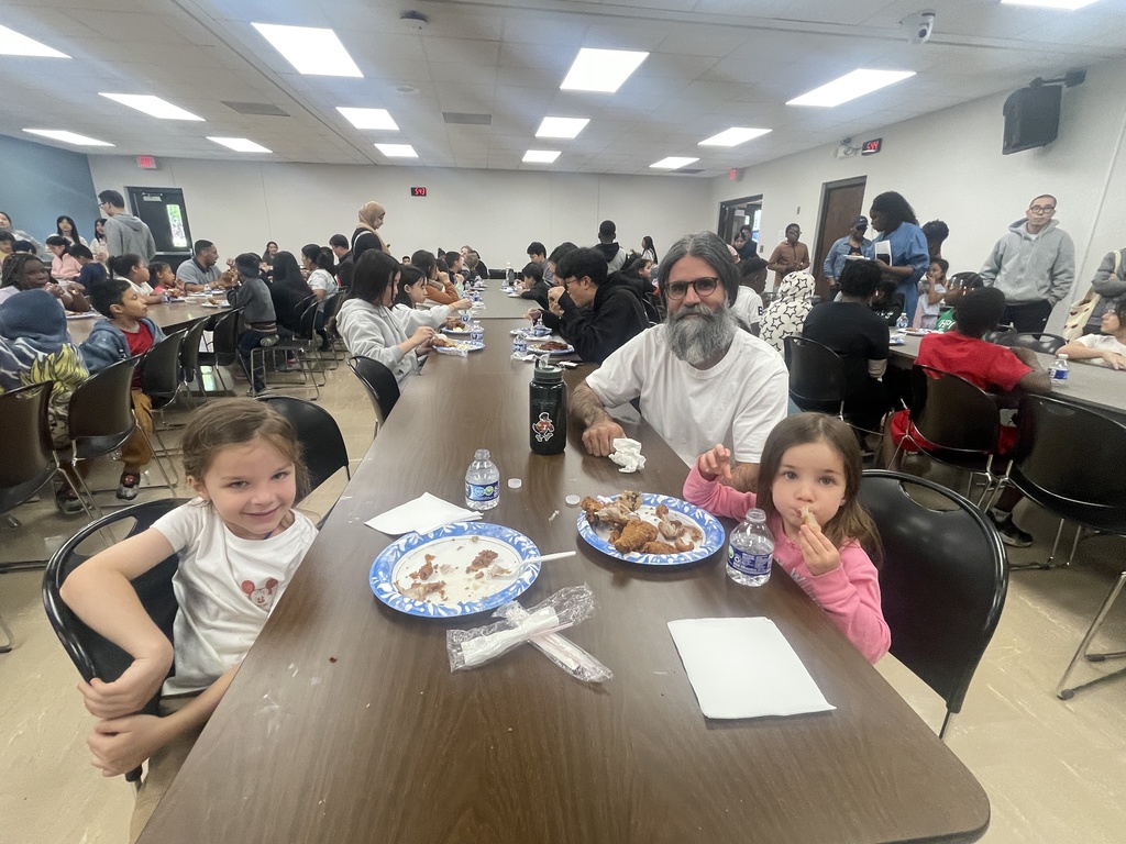 A table with three people, including two children, eating food. Several others sit at long tables in a cafeteria.
