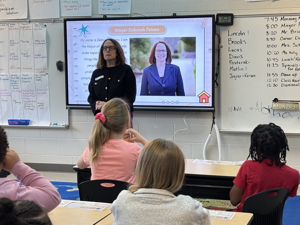 A teacher stands in front of a projector screen with a presentation, addressing children seated at desks.