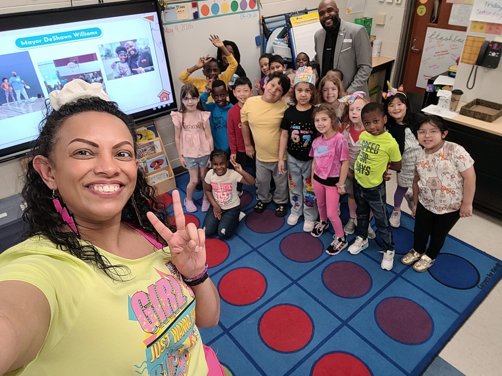 A woman takes a selfie with children in a classroom. The children stand on a colorful floor.
