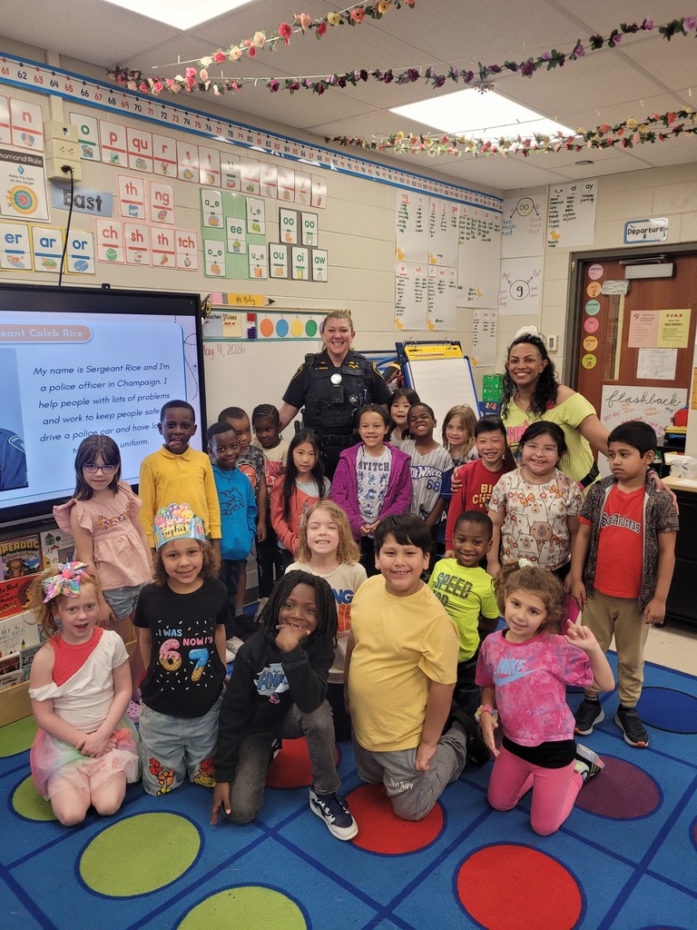 A group of children pose for a photo in a classroom, with a police officer standing behind them.