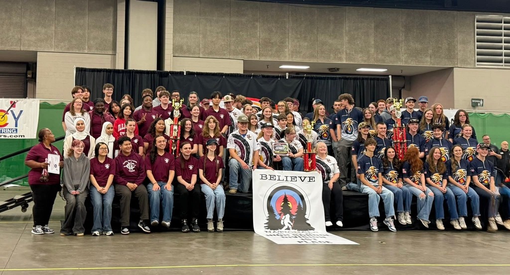 A group of people wearing matching maroon shirts, seated and standing on a stage, with a banner that reads "BELIEVE".