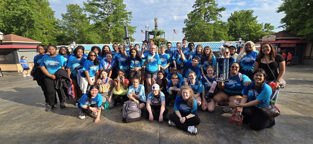 A group of people in matching blue shirts and various poses, with some holding a trophy. Background includes greenery and buildings.
